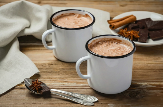Two rustic white mugs of healthy hot chocolate made with organic cacao powder, garnished with cinnamon sticks and star anise on a wooden table.