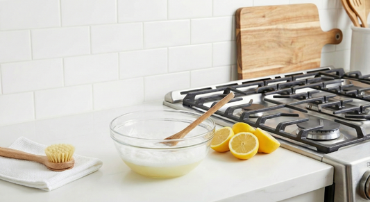 A glass bowl with a foaming baking soda and lemon solution and a wooden spoon sits on a white countertop next to halved lemons and a cleaning brush on a white towel, with a stovetop visible behind it